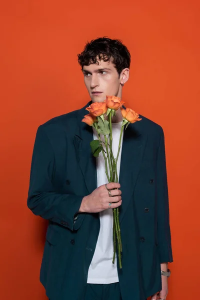 Модный молодой человек в блейзере держит оранжевые розы в течение 8 марта на красном фоне — стоковое фото