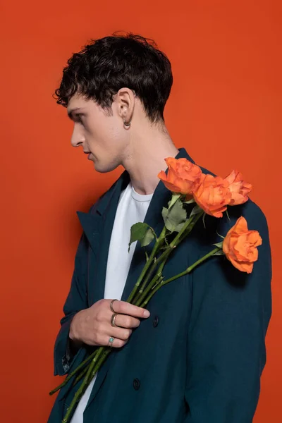 Vue latérale du jeune homme élégant en veste tenant des roses orange pour 8 mars sur fond rouge — Photo de stock