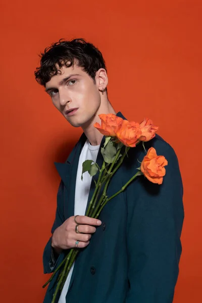 Trendy brunette man in blue suit holding orange flowers for 8 march on red background — Photo de stock