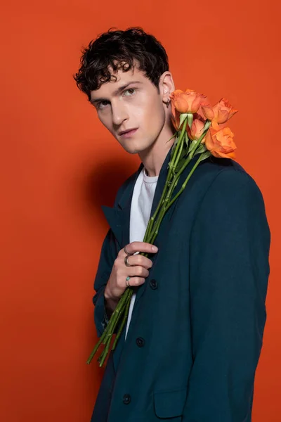 Portrait of stylish man in jacket holding roses near face on red background — Stock Photo