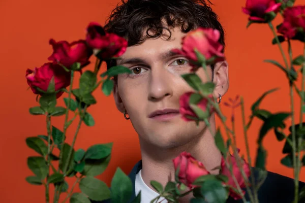 Curly homem olhando para a câmera perto de flores borradas no fundo vermelho — Fotografia de Stock