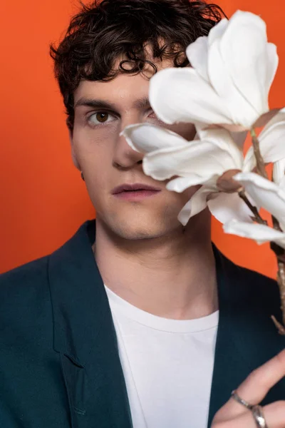 Curly man in jacket looking at camera near blurred magnolia flowers isolated on orange — Fotografia de Stock