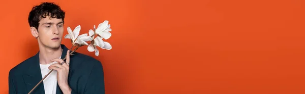 Brünetten Mann in Jacke Blick auf Magnolienzweig auf orangefarbenem Hintergrund, Banner — Stockfoto