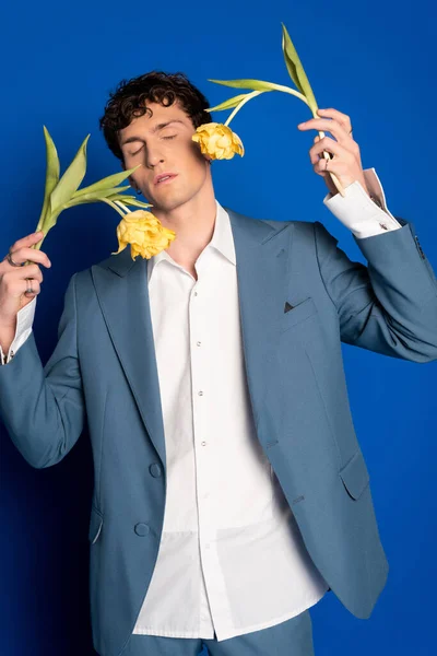 Trendy and curly young man holding tulips and closing eyes on blue background — Stock Photo