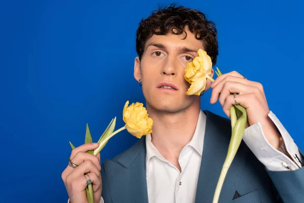 Portrait of curly man in jacket and shirt holding tulips near face isolated on blue — Stock Photo