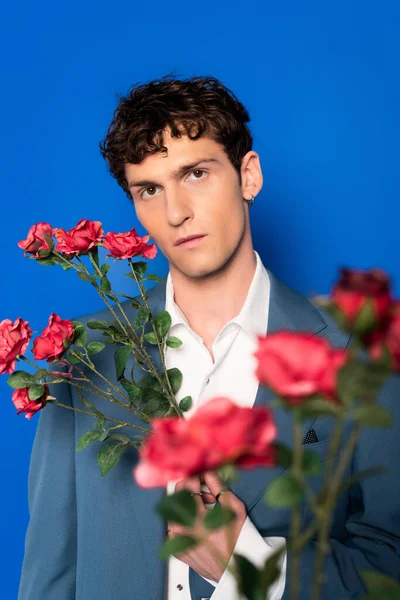 Портрет кучерявого молодого чоловіка в куртці позує з червоними квітами ізольовані на синьому — стокове фото