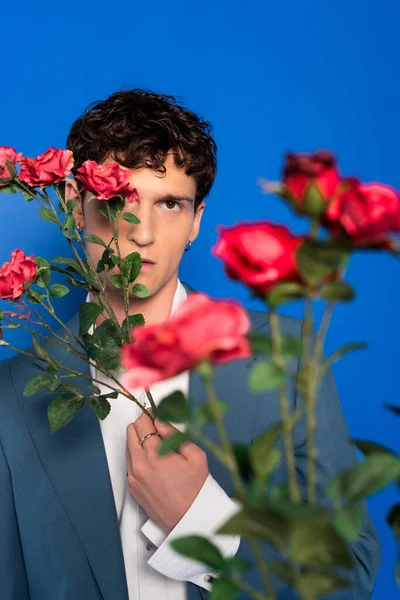 Curly man in stylish outfit covering face with flowers isolated on blue — Stock Photo