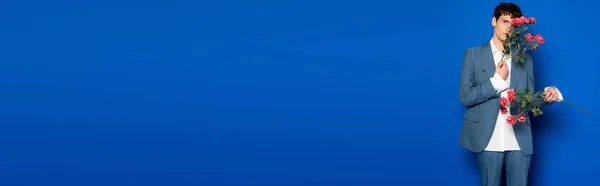 Young model in suit covering face with red roses on blue background, banner — стоковое фото