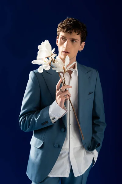 Modelo na moda em terno segurando flores de magnólia isoladas em azul marinho com luz solar — Fotografia de Stock