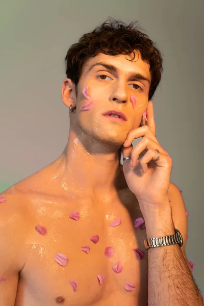Jeune homme avec des pétales sur le visage et la poitrine en regardant la caméra sur fond coloré — Photo de stock