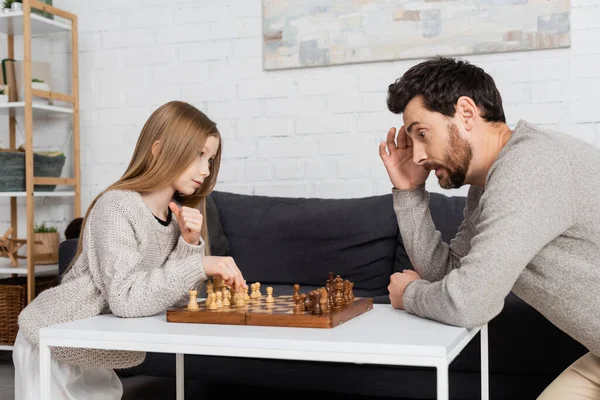 Thoughtful man touching head while playing chess with preteen daughter in living room — стоковое фото