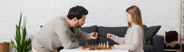 Side view of father and daughter holding figures while playing chess in living room, banner — Photo de stock