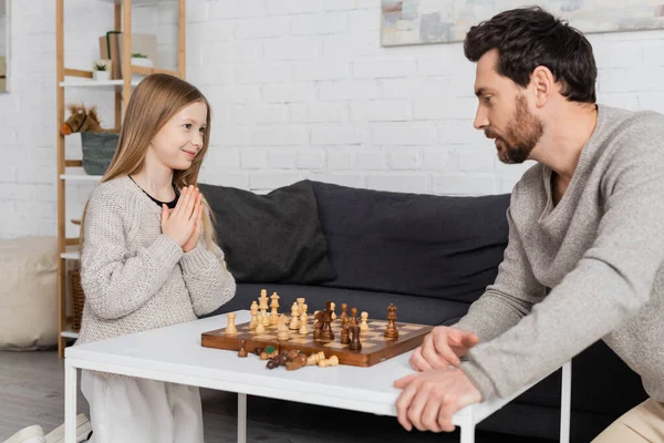 Fille souriante montrant s'il vous plaît geste tout en jouant aux échecs avec père à la maison — Photo de stock