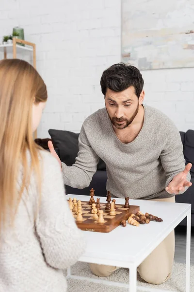 Confus homme barbu montrant geste haussant les épaules tout en jouant aux échecs avec fille préadolescente à la maison — Photo de stock