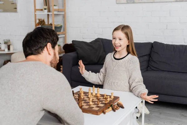 Gai preteen fille montrant wow geste près de père et échiquier sur table basse — Photo de stock
