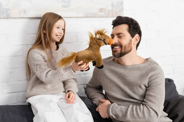 Cheerful girl showing toy horse to smiling dad sitting on couch with closed eyes — Foto stock