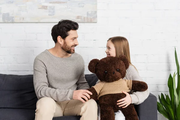 Happy girl holding soft toy and looking at cheerful bearded dad while sitting on sofa at home — Stock Photo