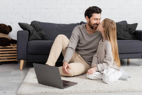 Cheerful father and daughter smiling at each other near laptop on floor in living room — Stock Photo