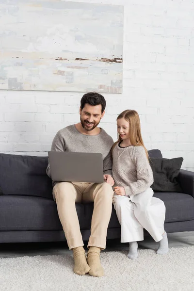 Joyful bearded man using laptop near smiling daughter on couch in living room — стоковое фото