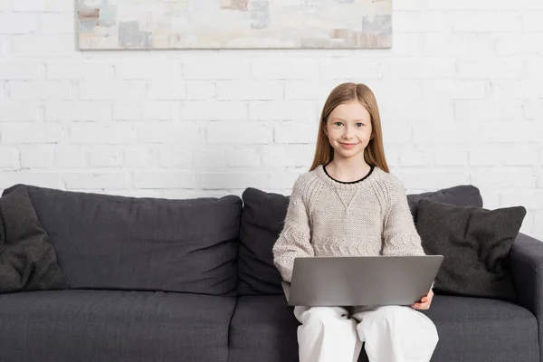 Pleased girl sitting on couch with laptop and smiling at camera — Stock Photo