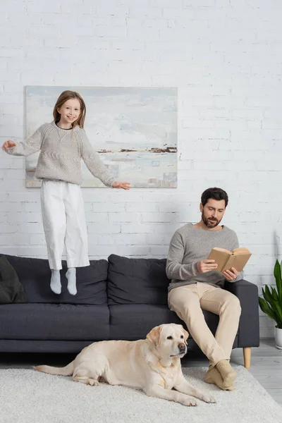 Cheerful girl levitating above couch near dad with book and labrador dog on carpet — Stockfoto