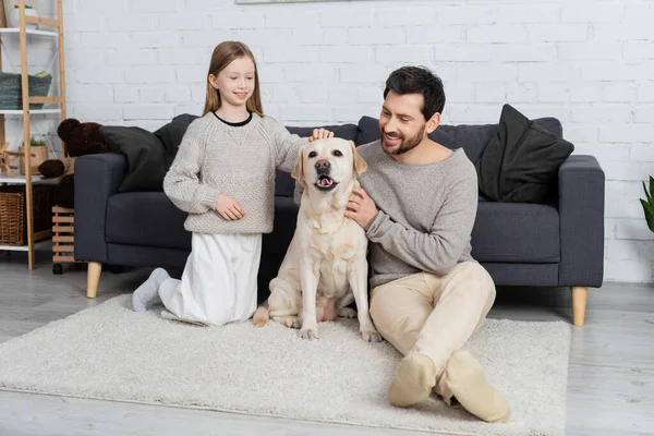 Joyful father and daughter petting labrador dog on carpet in living room — Stock Photo