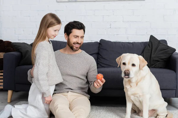Joyful bearded man embracing preteen daughter and holding ball near labrador dog at home — Stock Photo