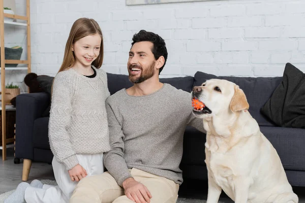 Bearded man with preteen girl smiling near labrador dog playing with ball in living room — Stock Photo