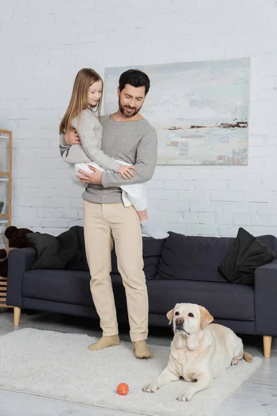 Full length of cheerful man holding daughter near labrador dog lying on carpet in living room — Stock Photo