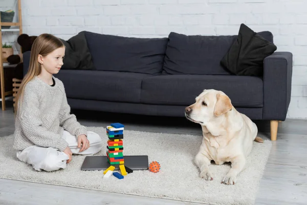 Happy girl and labrador dog looking at each other near wood blocks game and laptop on floor in living room — Stock Photo