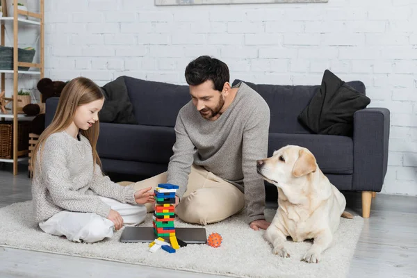 Smiling father and daughter playing wood blocks game near labrador dog on floor in living room — стоковое фото
