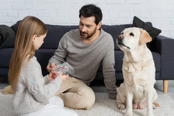 Girl holding palette with eye shadows while playing with dad near labrador dog on carpet in living room — Stock Photo