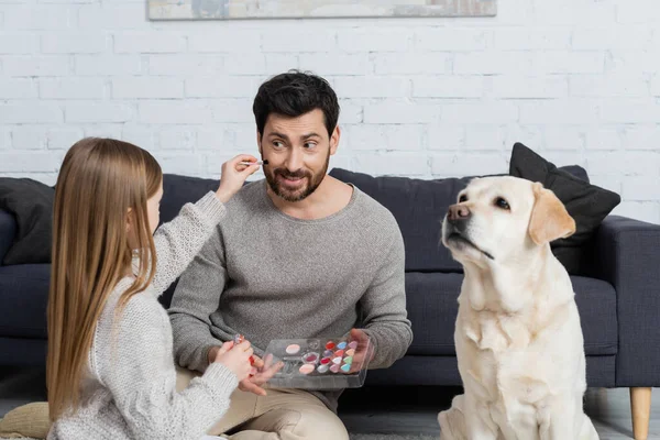 Preteen girl holding makeup palette while playing with smiling dad near labrador dog in living room — Stock Photo