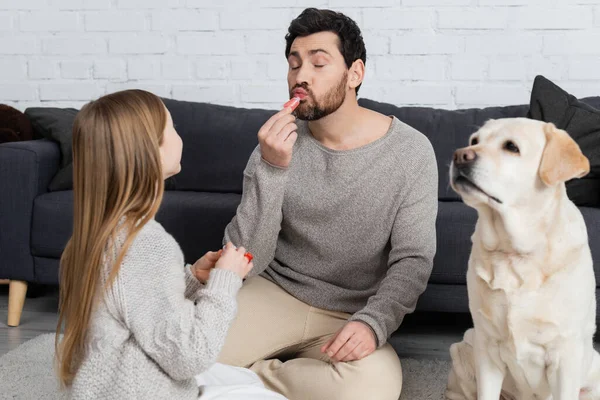 Bärtiger Mann mit geschlossenen Augen, der Lipgloss aufträgt, während er mit Tochter in der Nähe von Labrador-Hund im Wohnzimmer spielt — Stockfoto