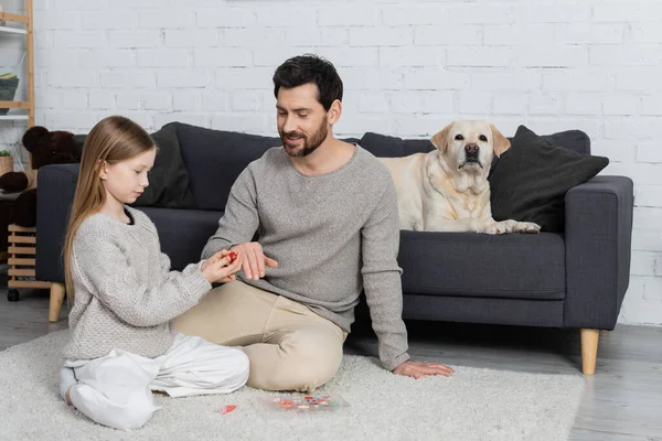 Preteen fille portant anneau sur doigt de père tout en jouant sur le sol près chien labrador couché sur le canapé dans le salon — Photo de stock