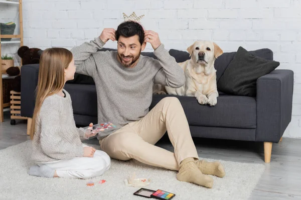 Cheerful bearded man wearing toy crown while sitting on floor near daughter and dog lying on sofa — Stock Photo