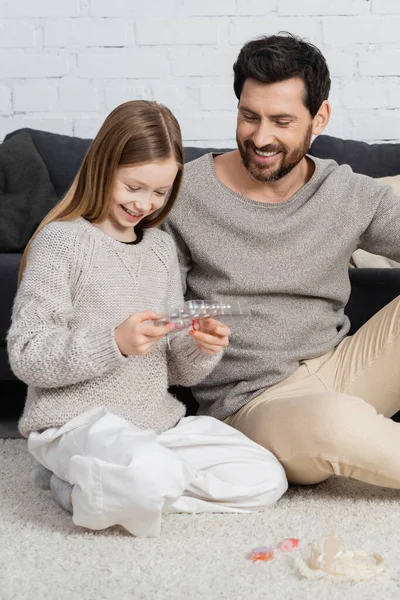 Feliz padre e hija mirando la paleta de maquillaje mientras están sentados en la alfombra cerca del sofá — Stock Photo