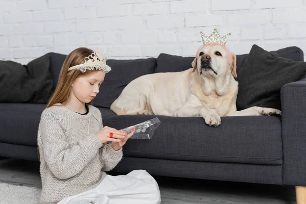 Preteen girl in crown looking at makeup palette while sitting near labrador on couch — Stock Photo