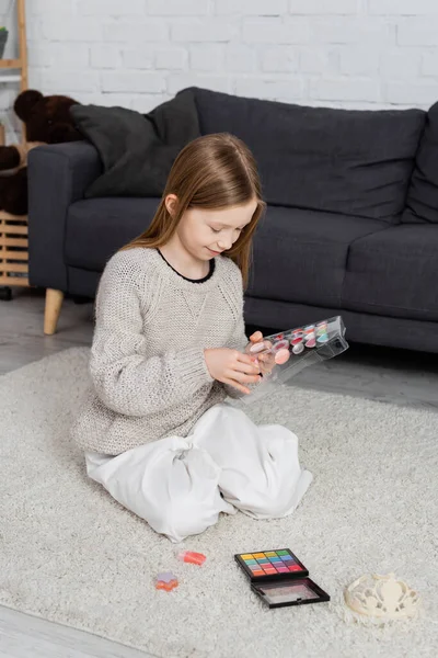 Felice preteen ragazza guardando tavolozza di trucco e seduto sul tappeto vicino divano — Foto stock