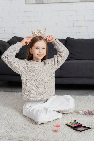 Happy preteen girl adjusting toy crown while sitting on carpet near makeup palette — Stock Photo