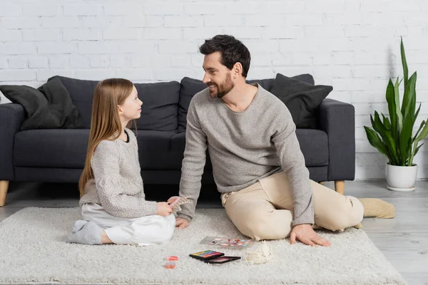 Happy father looking at smiling daughter while sitting on carpet near decorative cosmetics — Stock Photo