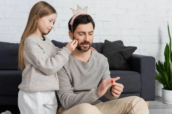 Girl holding cosmetic brush while applying makeup on cheek of smiling father in crown — Stock Photo