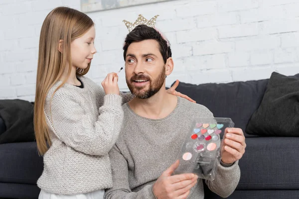 Girl holding cosmetic brush while applying makeup on face of smiling father in crown — Stock Photo
