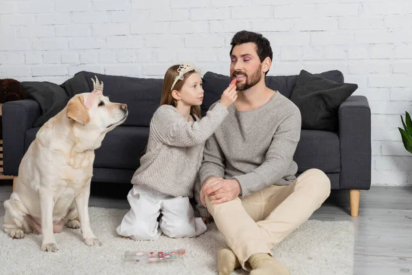 Happy girl in toy crown on head sitting near labrador and applying lip gloss on lips of father — Stock Photo