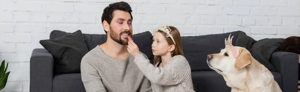 Happy girl in toy crown on head sitting near labrador and applying lip gloss on lips of father, banner — Stock Photo