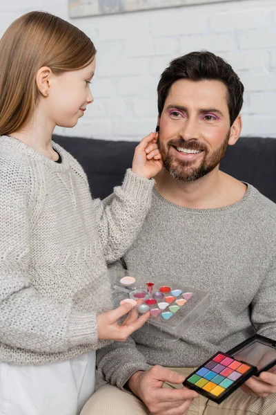 Preteen girl holding cosmetic brush while applying beauty product on cheek of bearded father — Stock Photo