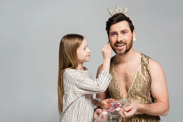 Happy kid applying decorative cosmetics on face of bearded father in crown and dress isolated on grey — Stock Photo