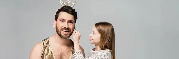Happy kid applying decorative cosmetics on face of bearded father in crown and dress isolated on grey, banner — Stock Photo