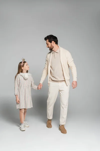Full length of happy father and daughter holding hands while looking at each other on grey — стоковое фото