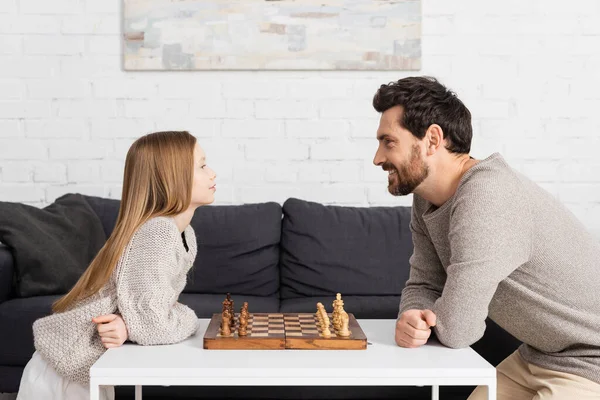 Vue latérale de la fille intelligente jouant aux échecs avec un père heureux dans le salon — Photo de stock